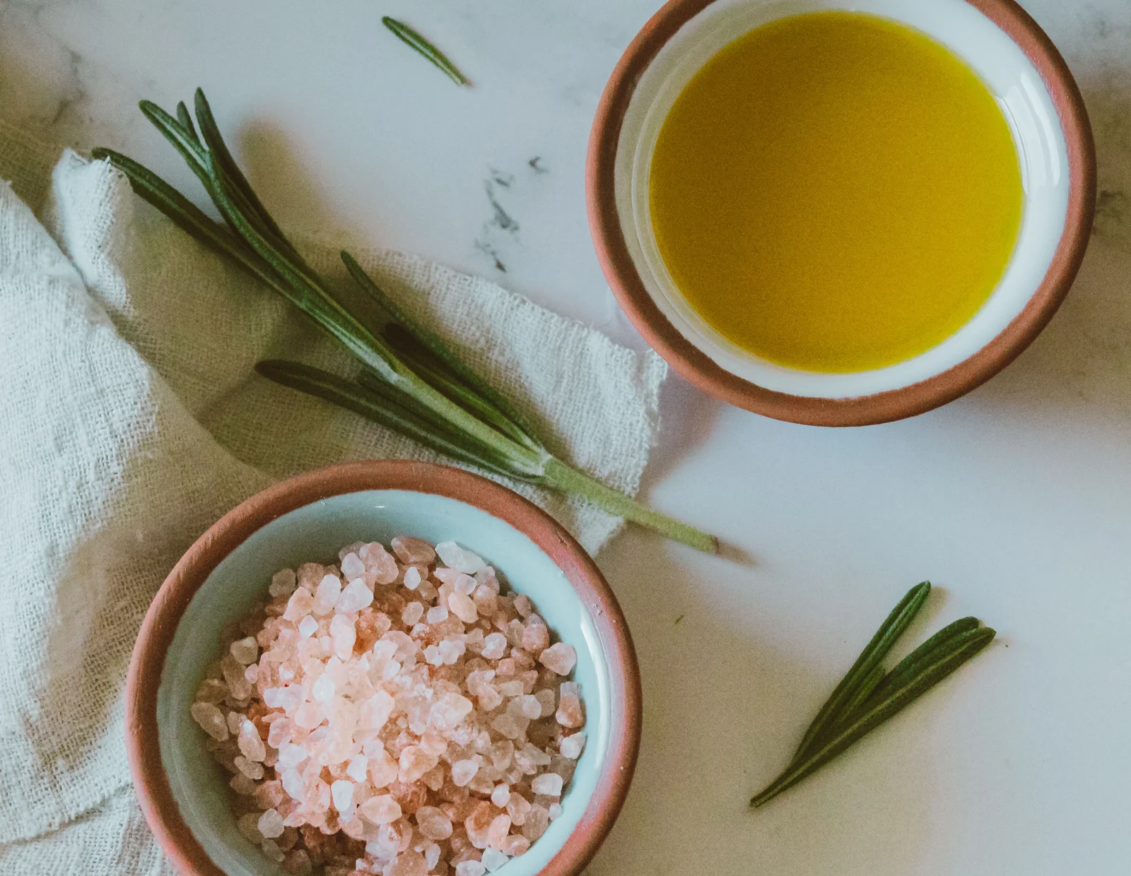 Bowls of olive oil and pink salt with rosemary on pale marble
